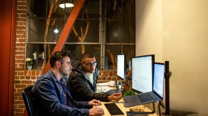 Real people, real offices. Two male developers working and collaborating in an casual enterprise office. They are using Visual Studio on a MacBook Pro with a multi-monitor set up. Code, develop, developer, engineer, Visual Studio, Azure.