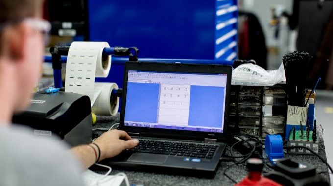 Mann sitzt an einem Schreibtisch, auf einem Bildschirm ist Code zu erkennen