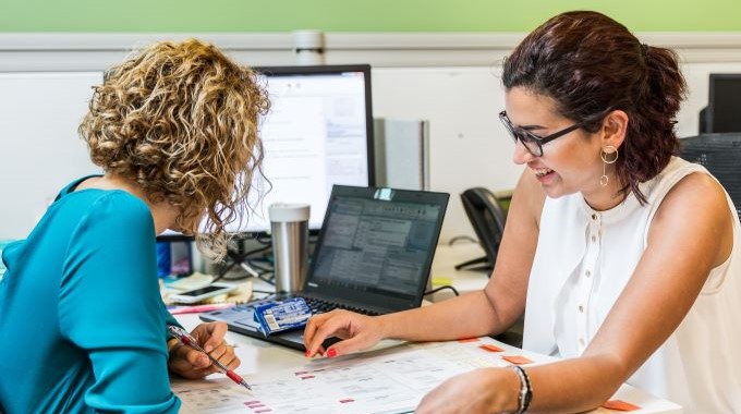 Zwei Frauen im Gespräch, sie sind über einen Schreibtisch gebeugt, auf dem Dokumente liegen und ein Laptop steht