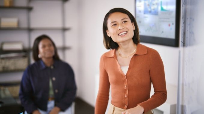 A woman holding a dry erase marker standing in front of a whiteboard smiling as she presents to another person sitting in the office behind her