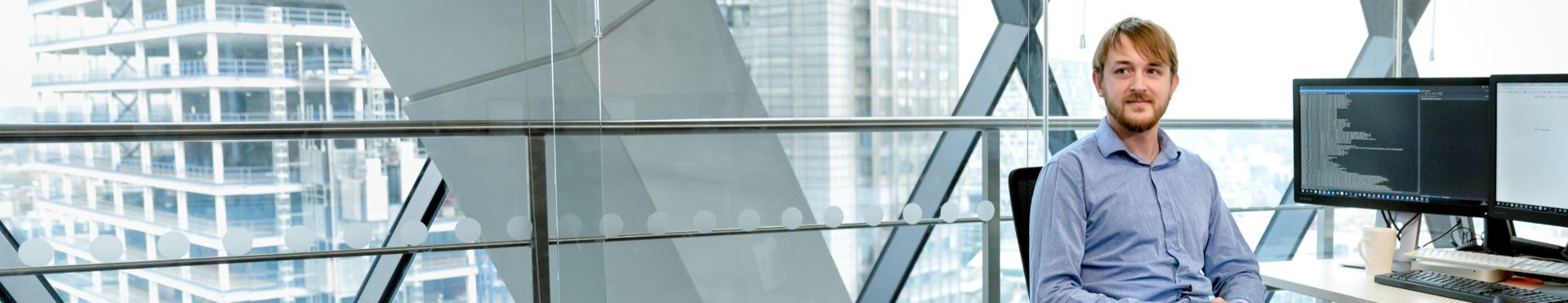 Portrait of male developer sitting at desk in open office. He is sitting with his legs crossed, looking off camera. Two monitors are on desk; one monitor‘s screen shows programming code.